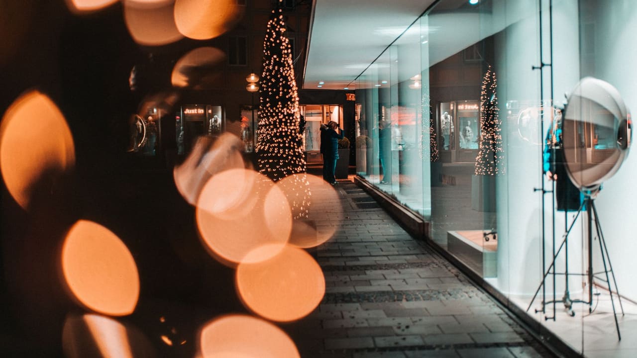 Christmas tree decorated with glowing garlands placed on paved sidewalk against modern store with glass walls at night