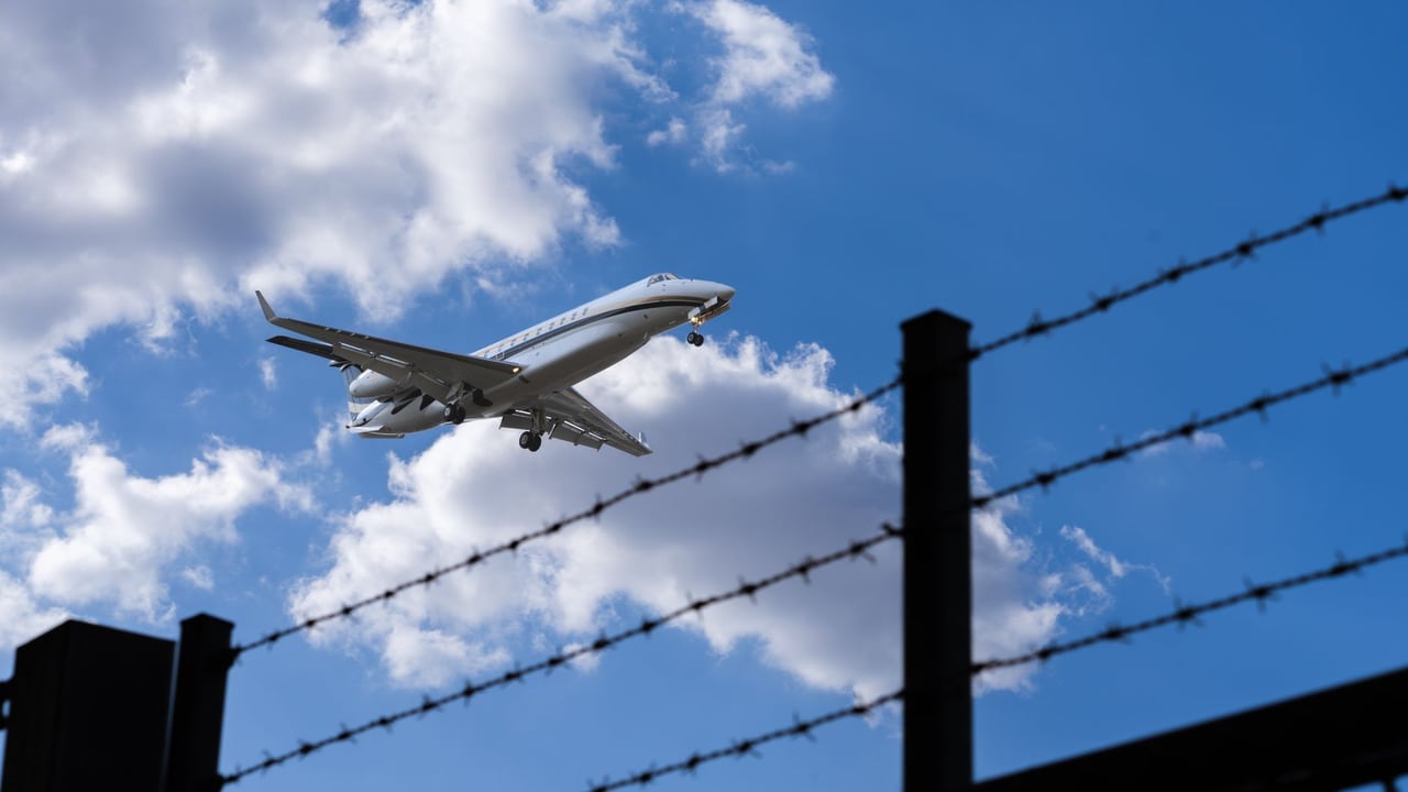 Jet approaching for landing, silhouetted against a bright blue sky with clouds and barbed wire in foreground.