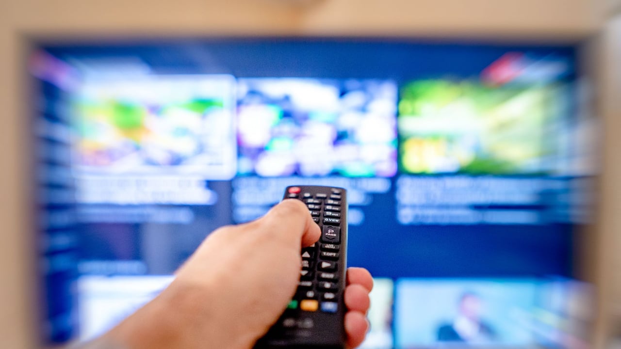 Hand holding remote control pointing at a blurred TV screen in a cozy indoor setting.