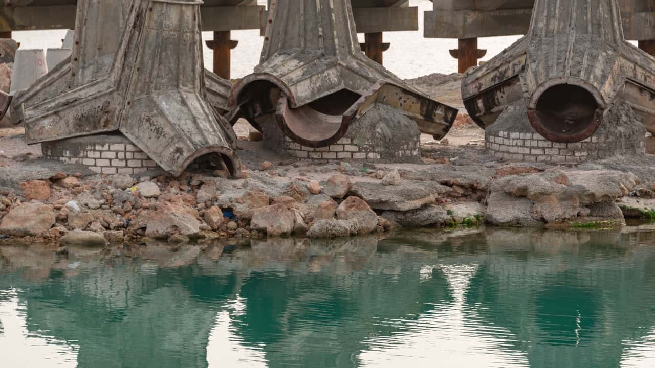 Concrete bridge construction site with rusty components and water reflection.