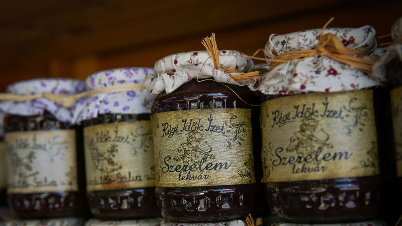 Close-up of rustic homemade jam jars with floral fabric covers and rustic labels on a wooden shelf.