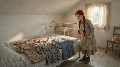 A red-haired girl in a plain apron stands by a bed where three simple dresses are laid out in a sunlit farmhouse bedroom.
