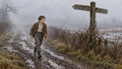A boy in a patched jacket walks along a foggy lane past a wet wooden signpost, hedges beaded with damp around him.