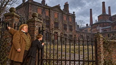 A boy looks through the bars of a courtyard gate at a dismal old brick house with barred and bricked-up windows beside a large brewery.