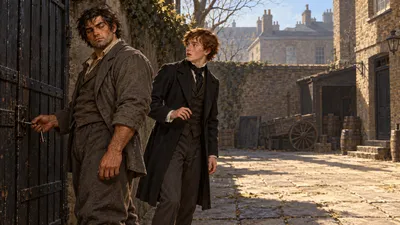 A burly slouching man in a plain grey suit turns from a locked gate holding a key, while a startled young man in a dark coat stands nearby in a bare courtyard.