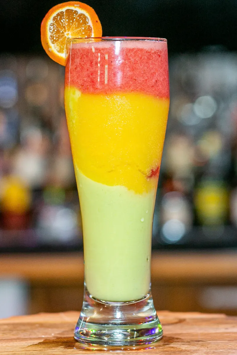 Vibrant layered smoothie in a tall glass, featuring bright red, yellow, and green hues, garnished with an orange slice.