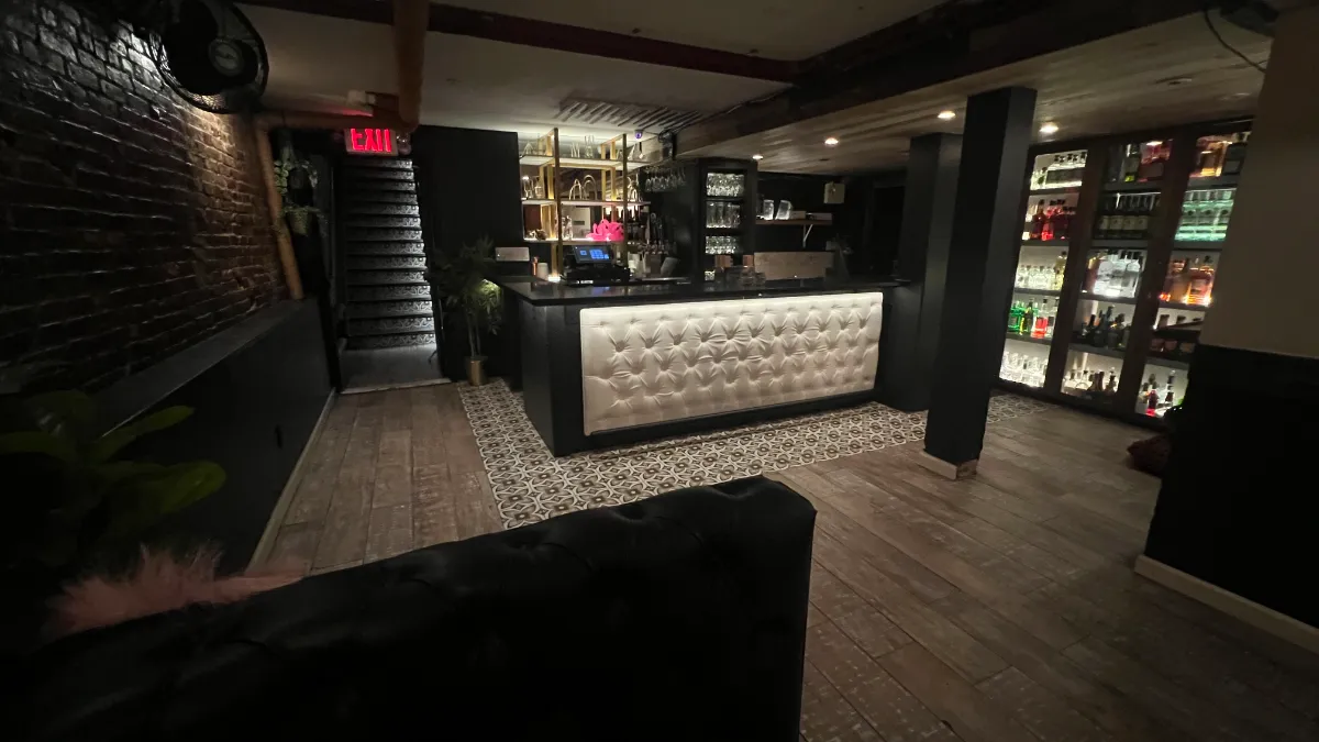 A stylish interior view of the Loreley Beer Garden bar, featuring a sleek white tufted bar counter illuminated with soft lighting, surrounded by an array of liquor bottles displayed on illuminated shelves against a dark brick backdrop. The space is ideal for gatherings and special events.