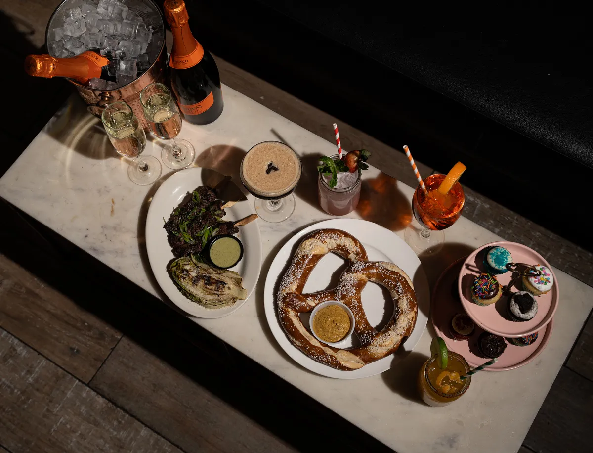 An enticing spread featuring a warm pretzel with dipping sauce, gourmet grilled greens, and an array of colorful desserts including cupcakes, complemented by a selection of cocktails and sparkling beverages at Loreley Beer Garden in the Lower East Side, NY 10002.