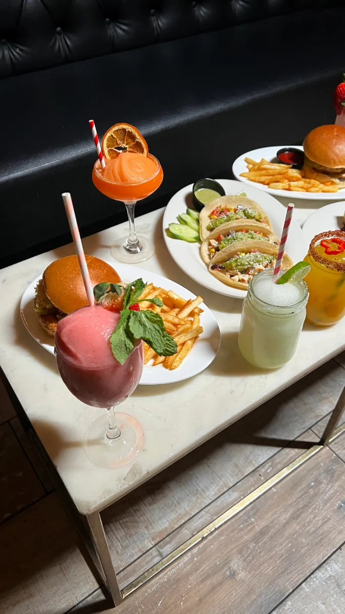 A vibrant display of cocktails and delicious food at Loreley Beer Garden in NYC. The image features a refreshing strawberry cocktail garnished with mint, a bright orange drink topped with an orange slice, and a classic margarita. Accompanying the drinks are crispy French fries, a juicy burger, and a
