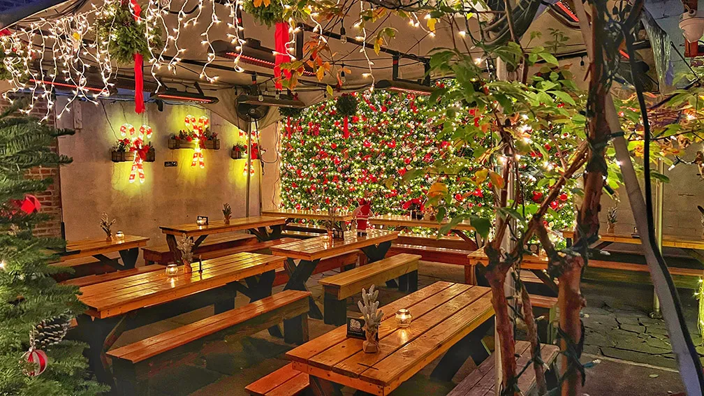 A beautifully decorated outdoor seating area at Loreley Beer Garden, featuring wooden picnic tables, twinkling fairy lights, and a vibrant holiday-themed wall adorned with festive greenery and ornaments. Perfect for gatherings and celebrations in the Lower East Side, NY 10002.