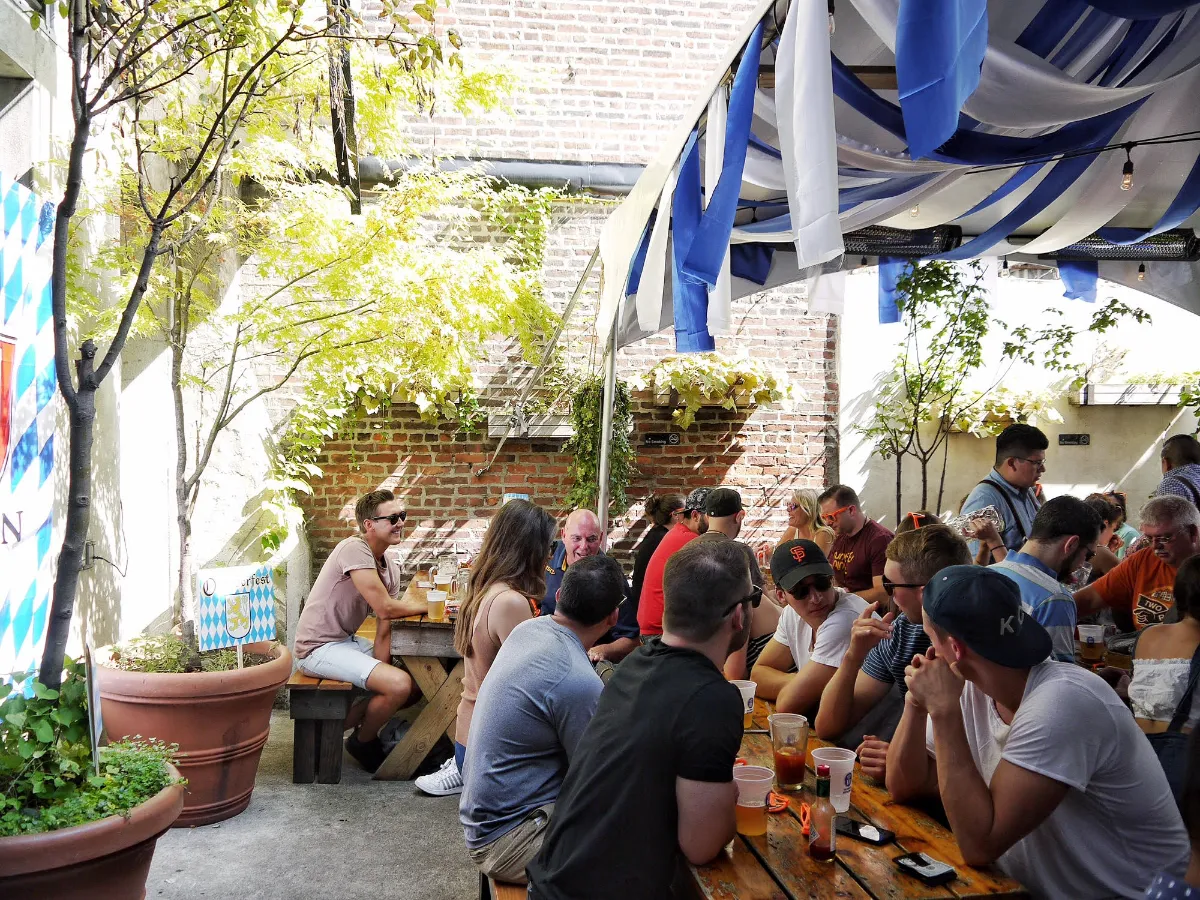 A lively scene at Loreley Beer Garden in NYCs Lower East Side, with patrons enjoying food and drinks at communal wooden tables under a festive canopy. The outdoor space, adorned with greenery and flag decorations, is bustling with people socializing, making it an ideal spot for gatherings and events