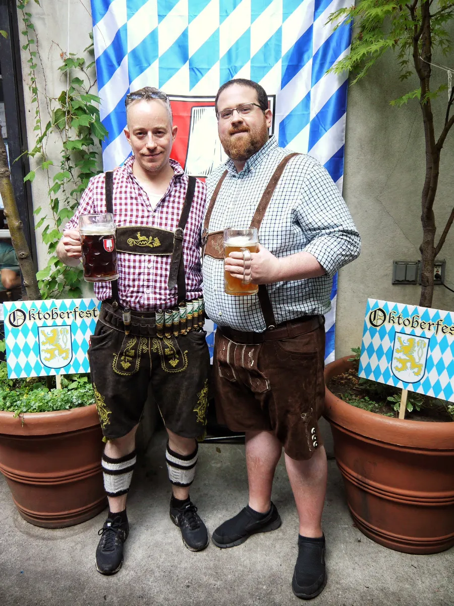 Two men in traditional Oktoberfest attire stand together, each holding a large mug of beer. They are in front of a blue and white checkered backdrop with an Oktoberfest sign, surrounded by potted plants. The setting exudes a festive atmosphere, perfect for enjoying German beer and celebrating at events like