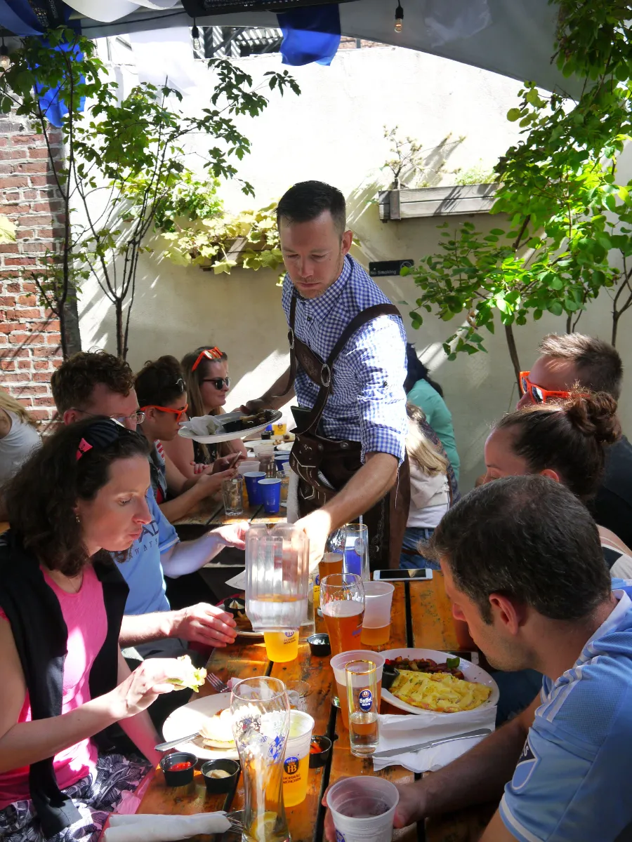 A lively scene at Loreley Beer Garden in NYCs Lower East Side (10002) featuring patrons enjoying delicious German food and craft beers. A waiter, dressed in traditional attire, pours water for guests seated at a wooden table adorned with plates of food, drinks in vibrant glasses, and a cheerful atmosphere