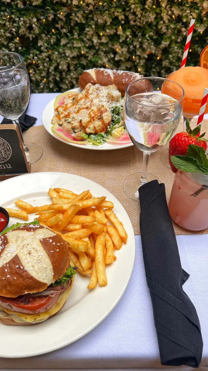 A mouthwatering meal at Loreley Beer Garden featuring a juicy burger with a pretzel bun, served with crispy fries and a side of dipping sauce, alongside a fresh salad and a specialty cocktail adorned with a strawberry and mint garnish, set against a backdrop of lush greenery, perfect for dining