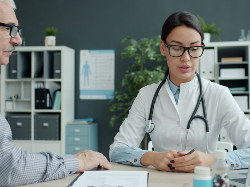 Caring female doctor is talking to elderly man patient holding hand expressing care in clinic. Healthcare, people and communication concept.