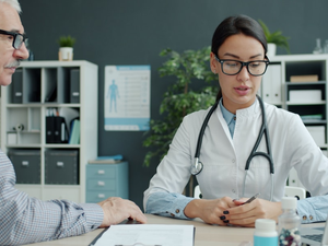 Caring female doctor is talking to elderly man patient holding hand expressing care in clinic. Healthcare, people and communication concept.