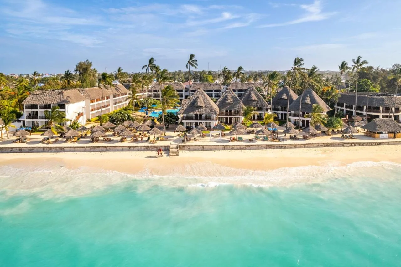 Aerial drone view of thatched resort buildings along a white-sand beach.