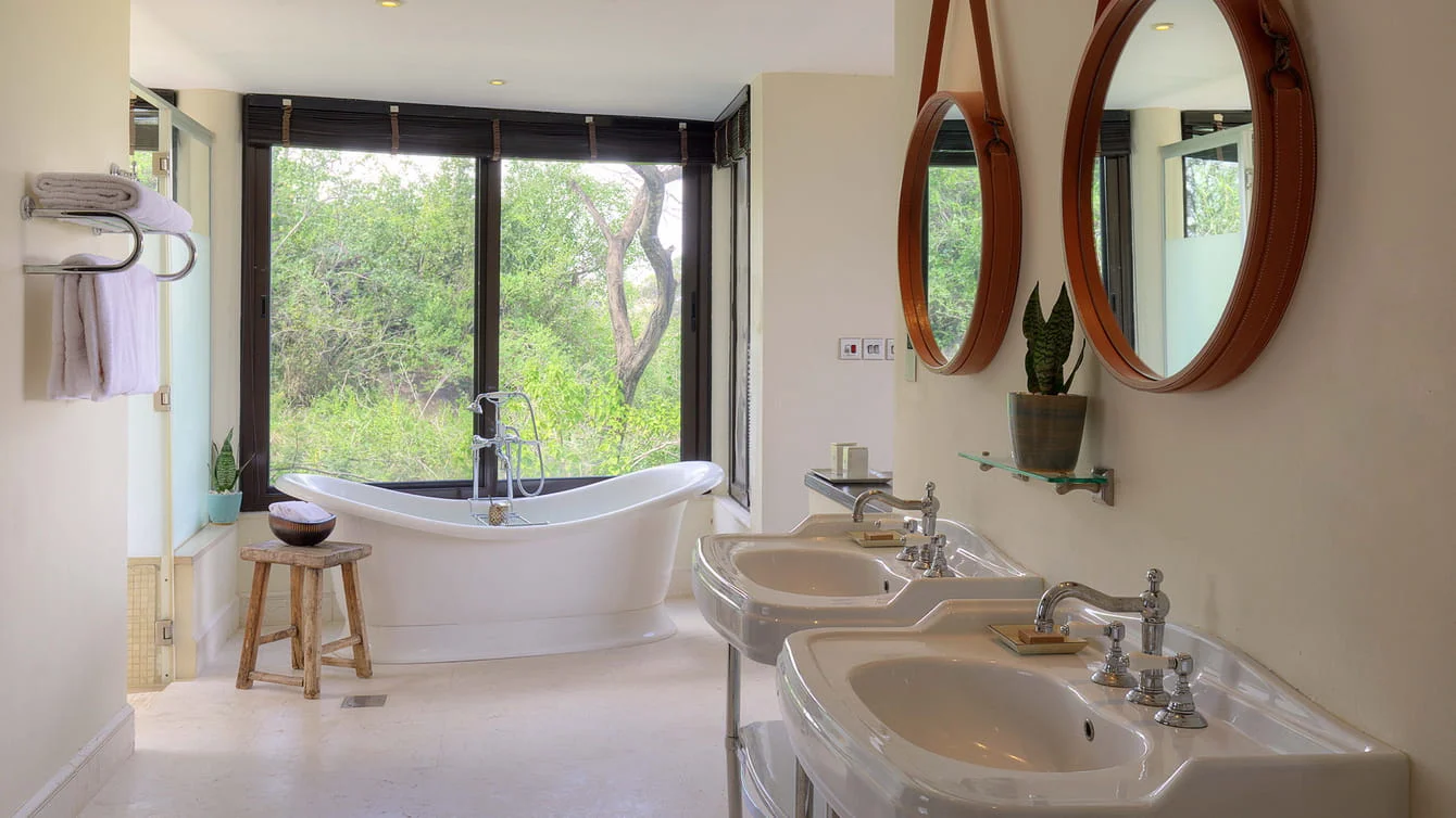 En-suite bathroom featuring double vessel sinks, round wall mirrors, and a deep bathtub overlooking nature.