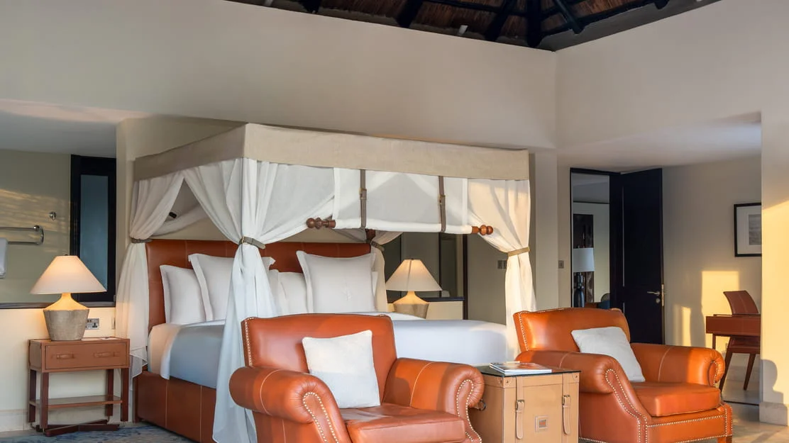 Canopy king bed wrapped in white netting with leather armchairs in a luxury Serengeti suite bedroom.