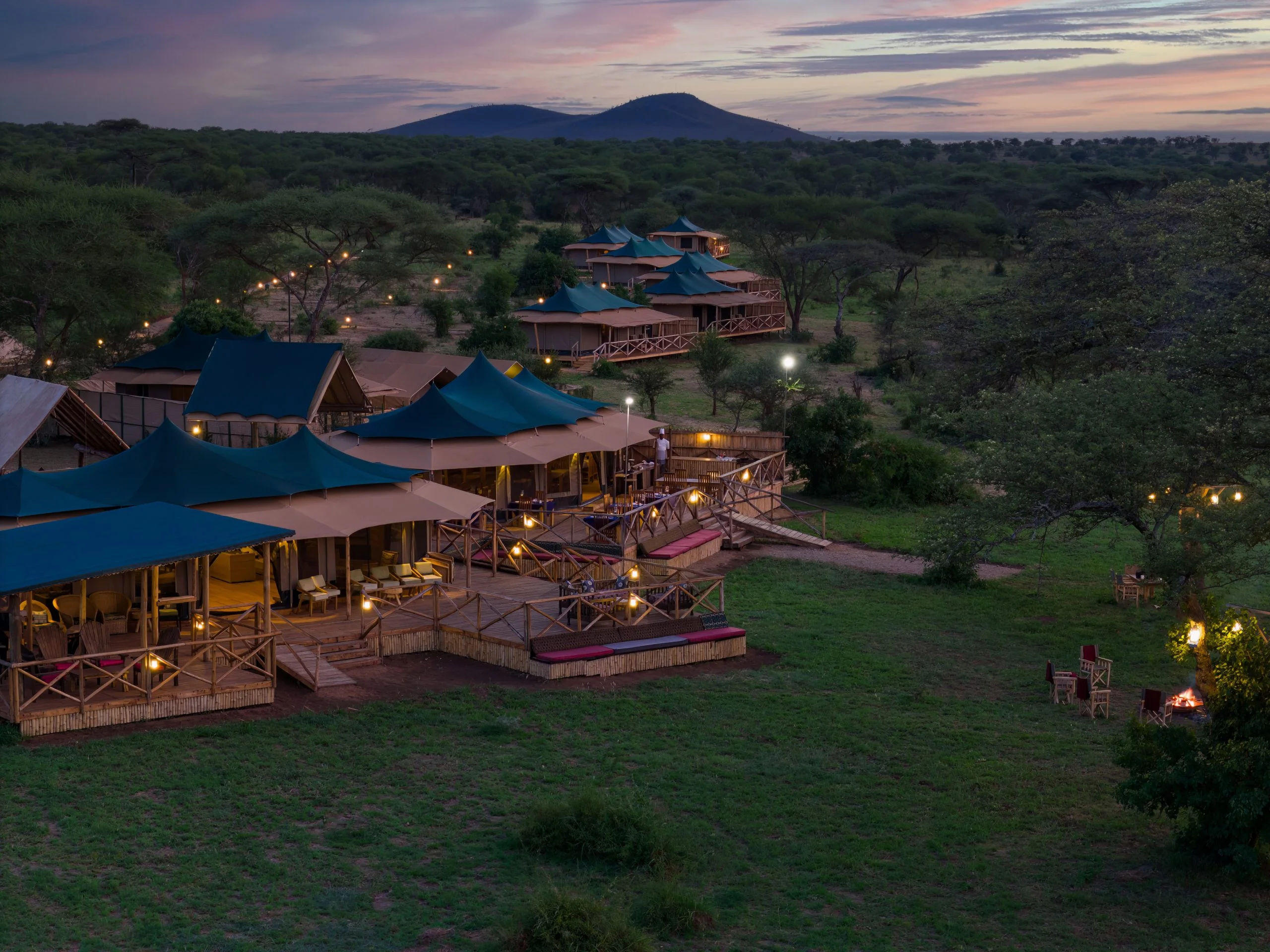 Aerial twilight view of lit safari tents and deck fire pit surrounded by Serengeti landscape.