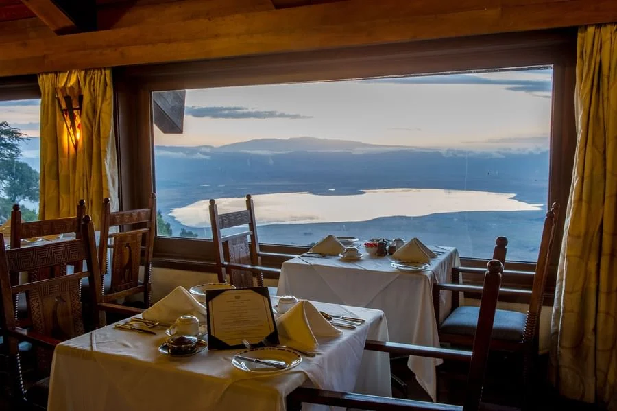 Dining table set with white linen next to large windows overlooking panoramic views of Ngorongoro Crater.
