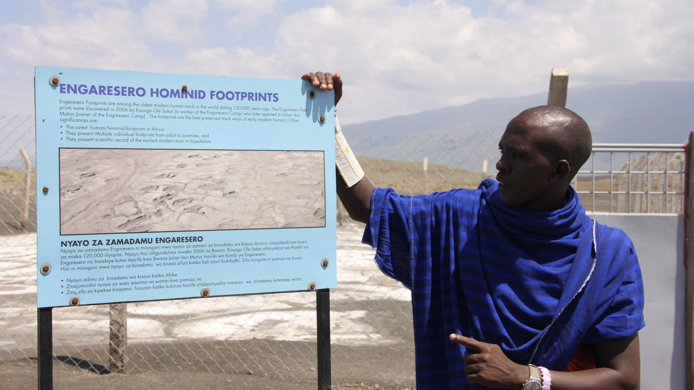 Maasai guide standing in traditional shúkà beside the Engaresero Hominid Footprints info sign, Tanzania.