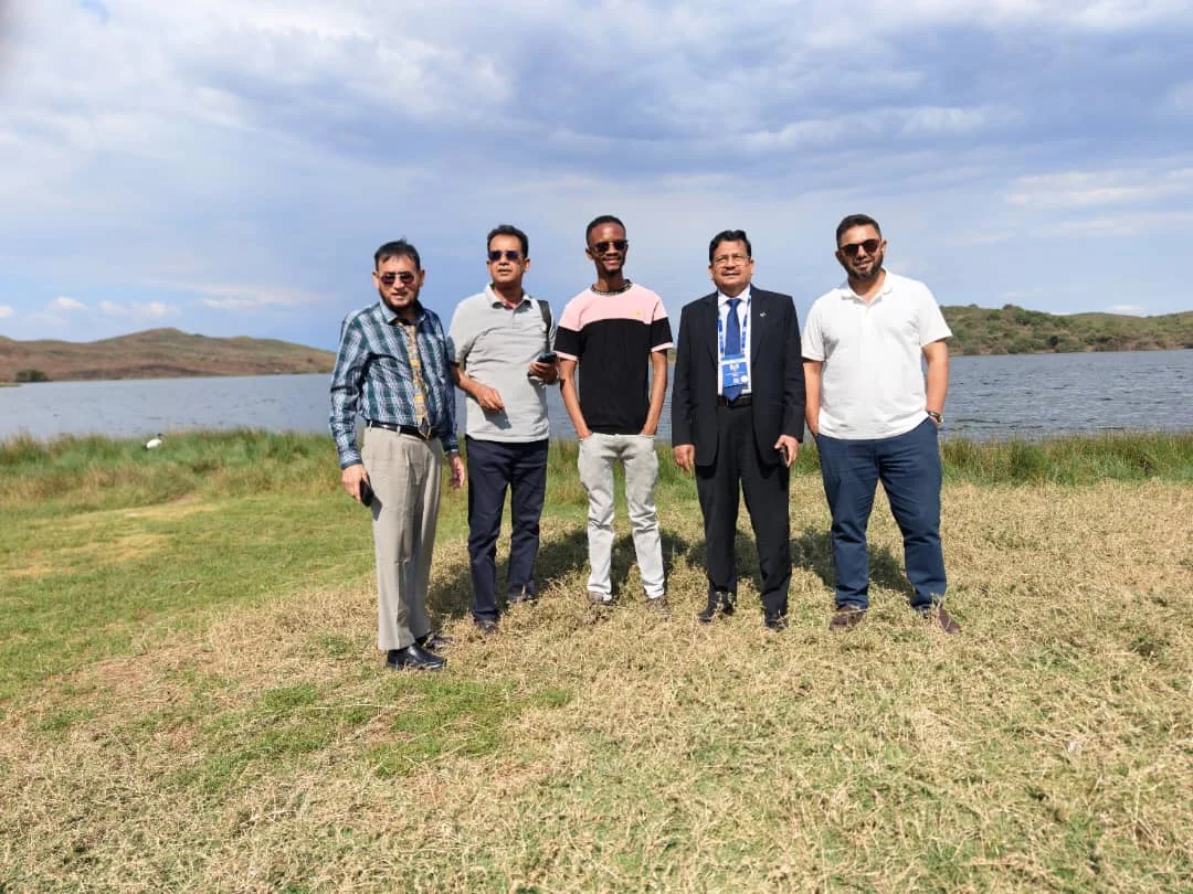 Group of five men standing in a row on a grassy shore next to a lake.