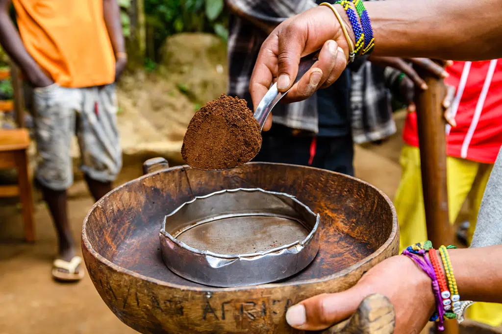 Hands filtering roasted ground coffee powder into a traditional metal sieve during a cultural coffee tour