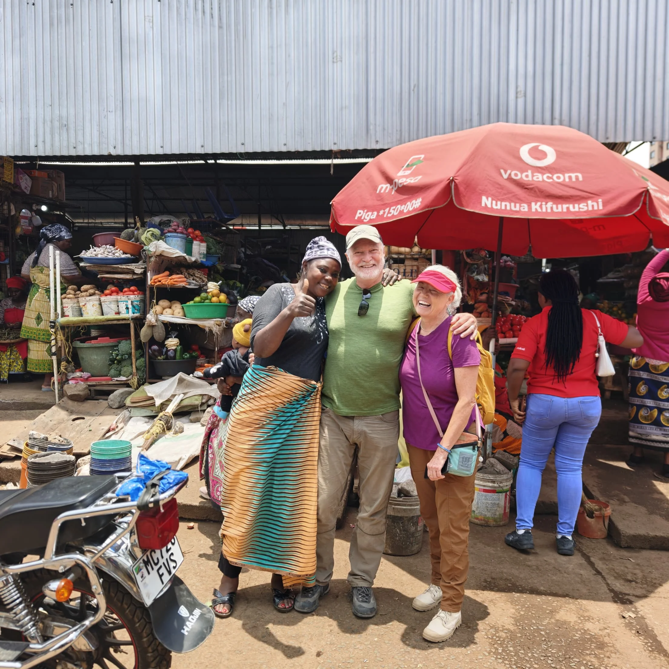 Tourists posing with a local vendor under a red umbrella at a market stall in Arusha Central Market