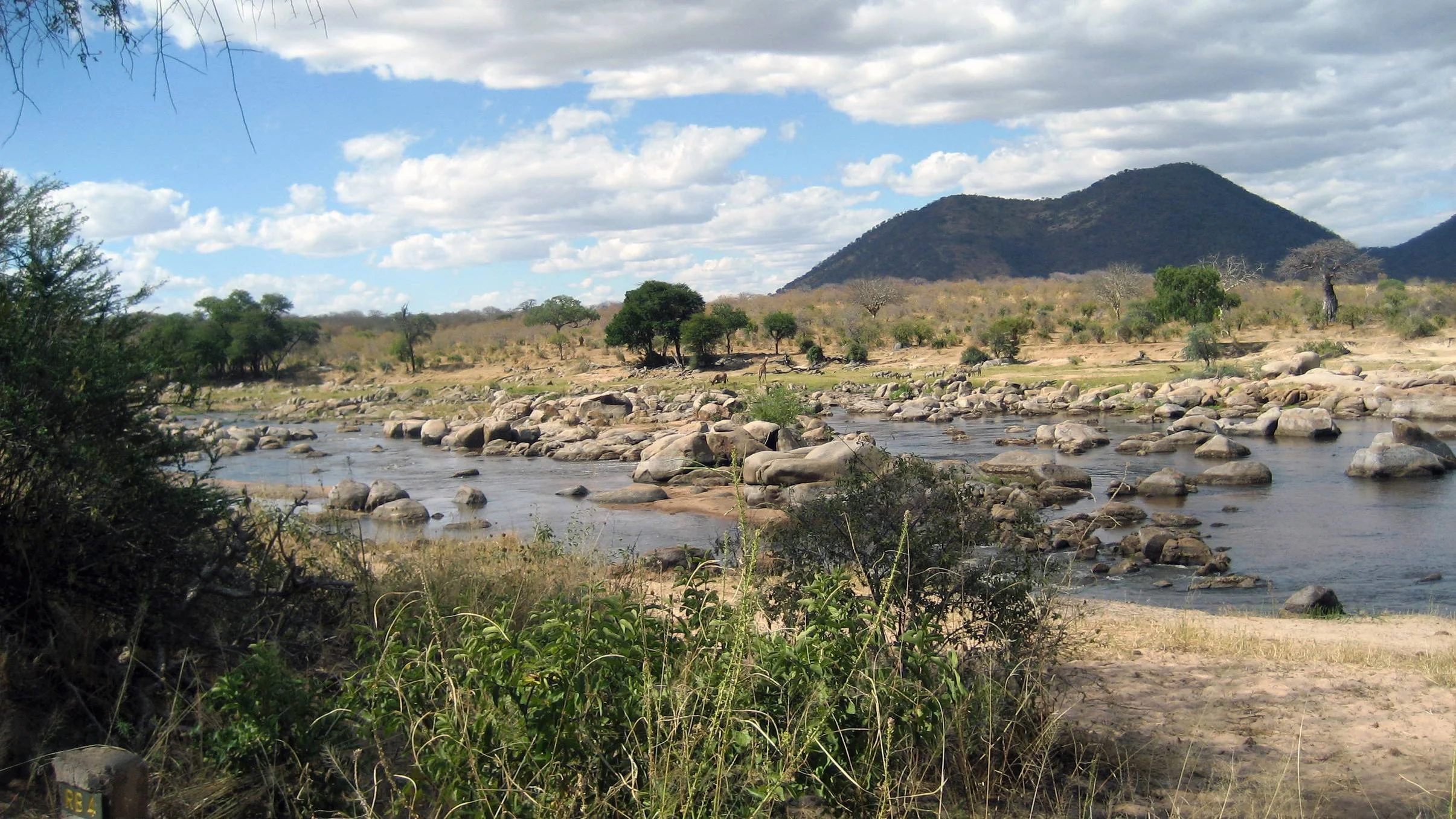 A rocky section of the Ruaha River under a cloudy blue sky with distant mountains.