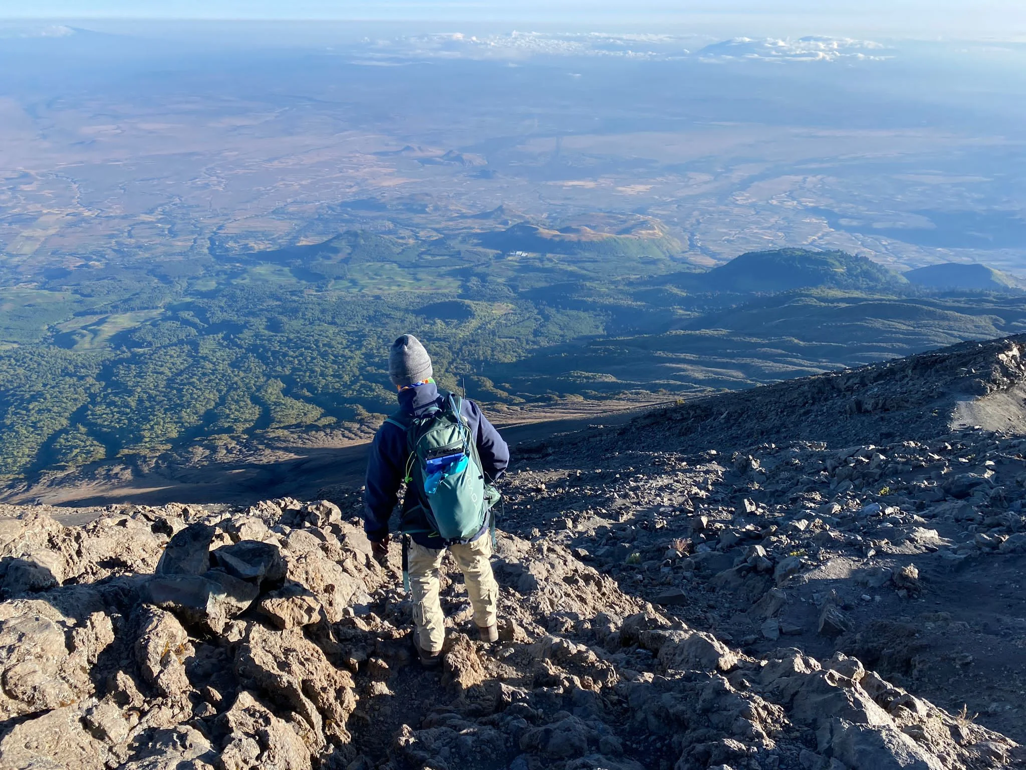Hiker with backpack standing on rocky ridge overlooking vast valley plains below Mount Kilimanjaro, Tanzania.
