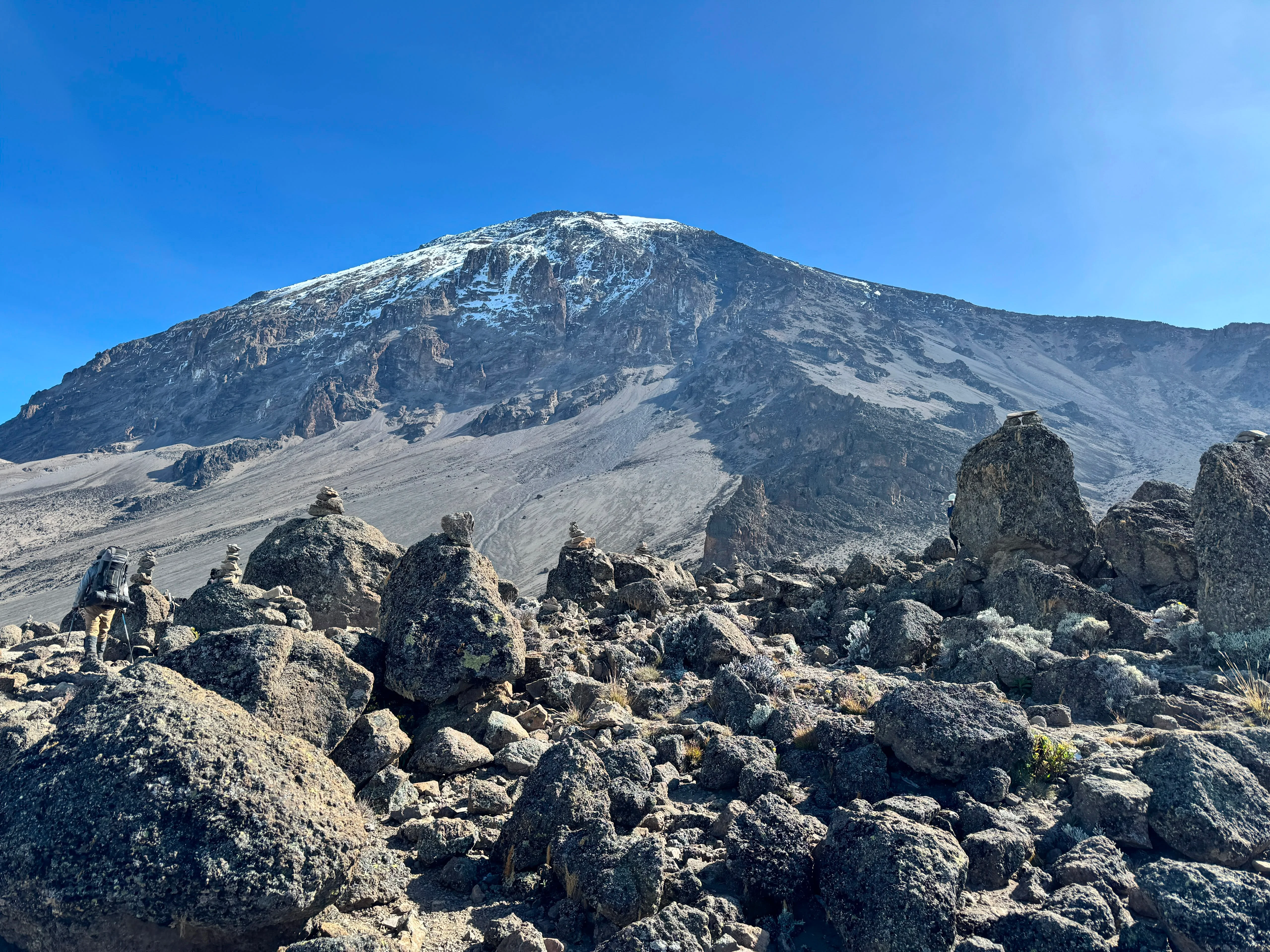 Rocky volcanic terrain leads toward Mount Kilimanjaro's snow-dusted summit beneath a bright blue sky along the Machame Route.