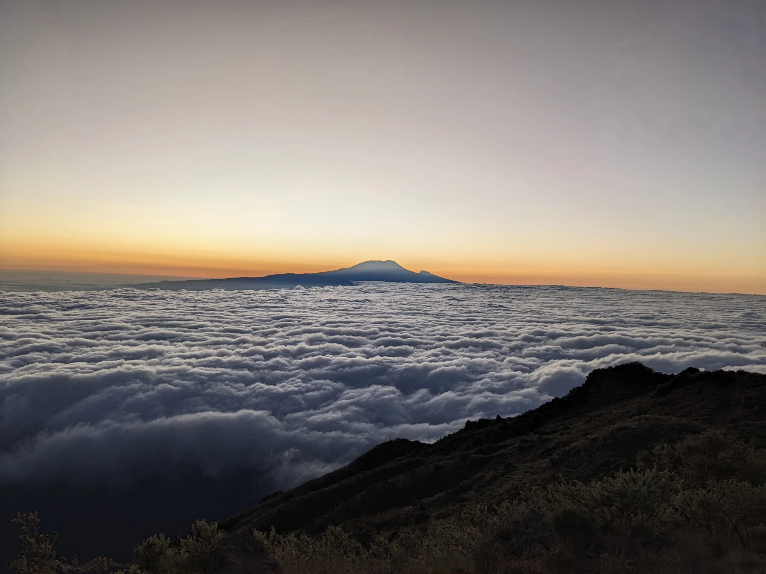 Sunrise above a sea of clouds on Mount Kilimanjaro, with distant Mawenzi Peak silhouetted against a warm morning horizon sky.