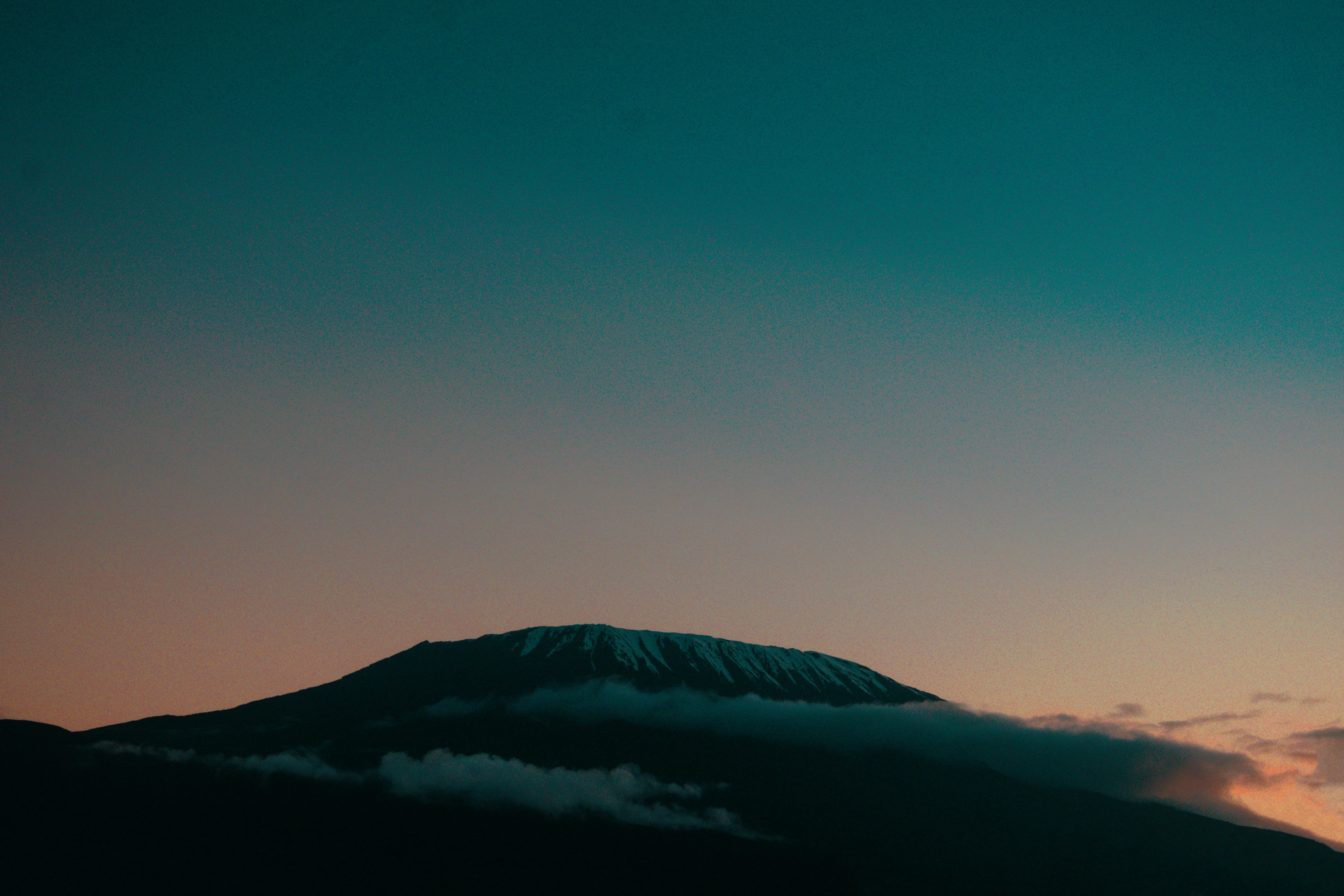 Mount Kilimanjaro appears in silhouette at sunset, with its snowy summit rising above clouds beneath a dramatic Tanzania sky.