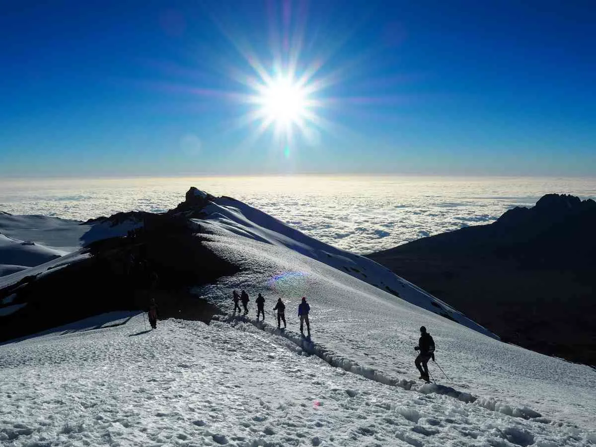 Trekkers navigating snowy summit slopes under a bright sun above clouds on Mount Kilimanjaro, Tanzania.