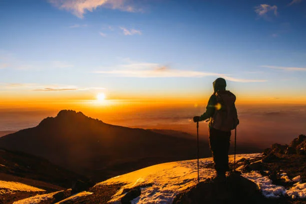 Silhouette of a trekker with walking poles at sunrise on a rocky mountain summit trail on Kilimanjaro.