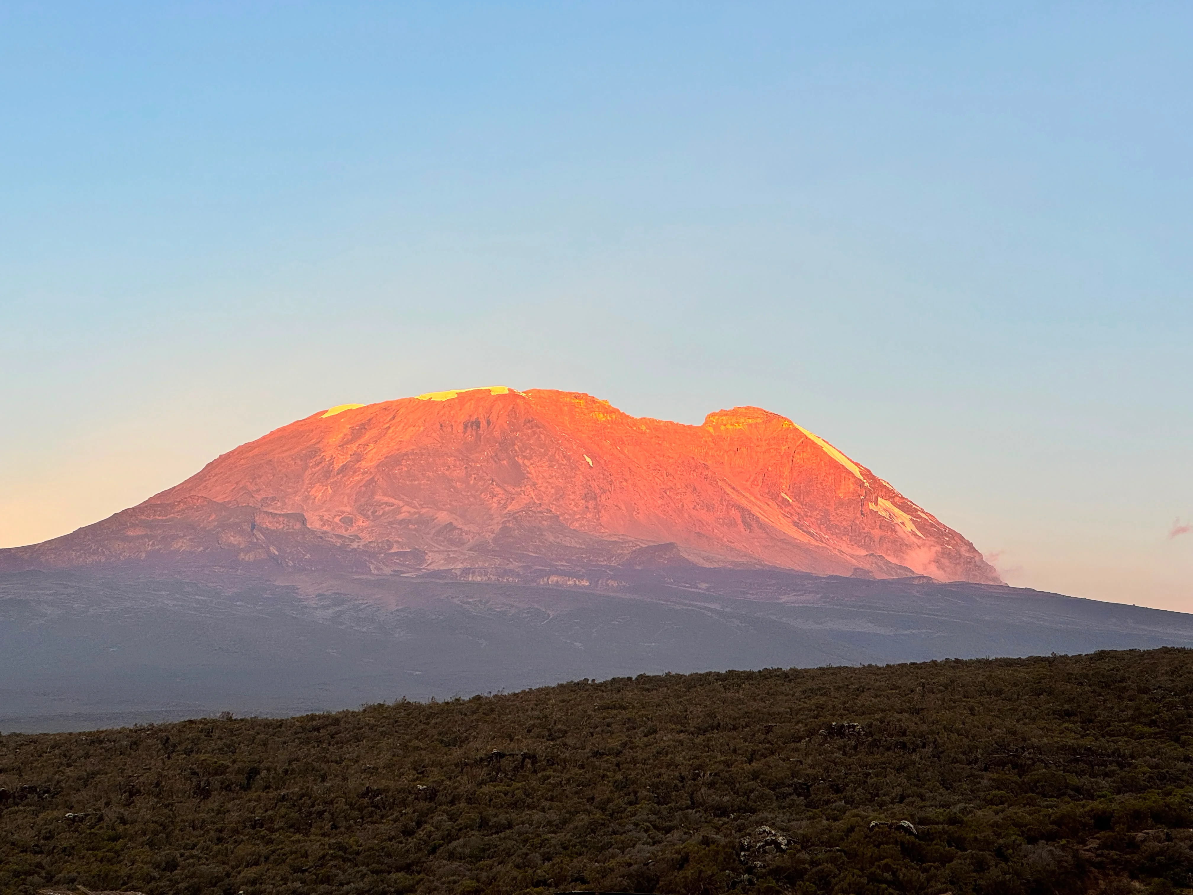 Mount Kilimanjaro glows orange at sunrise above dark forested slopes, creating a dramatic view along Tanzania's Lemosho Route