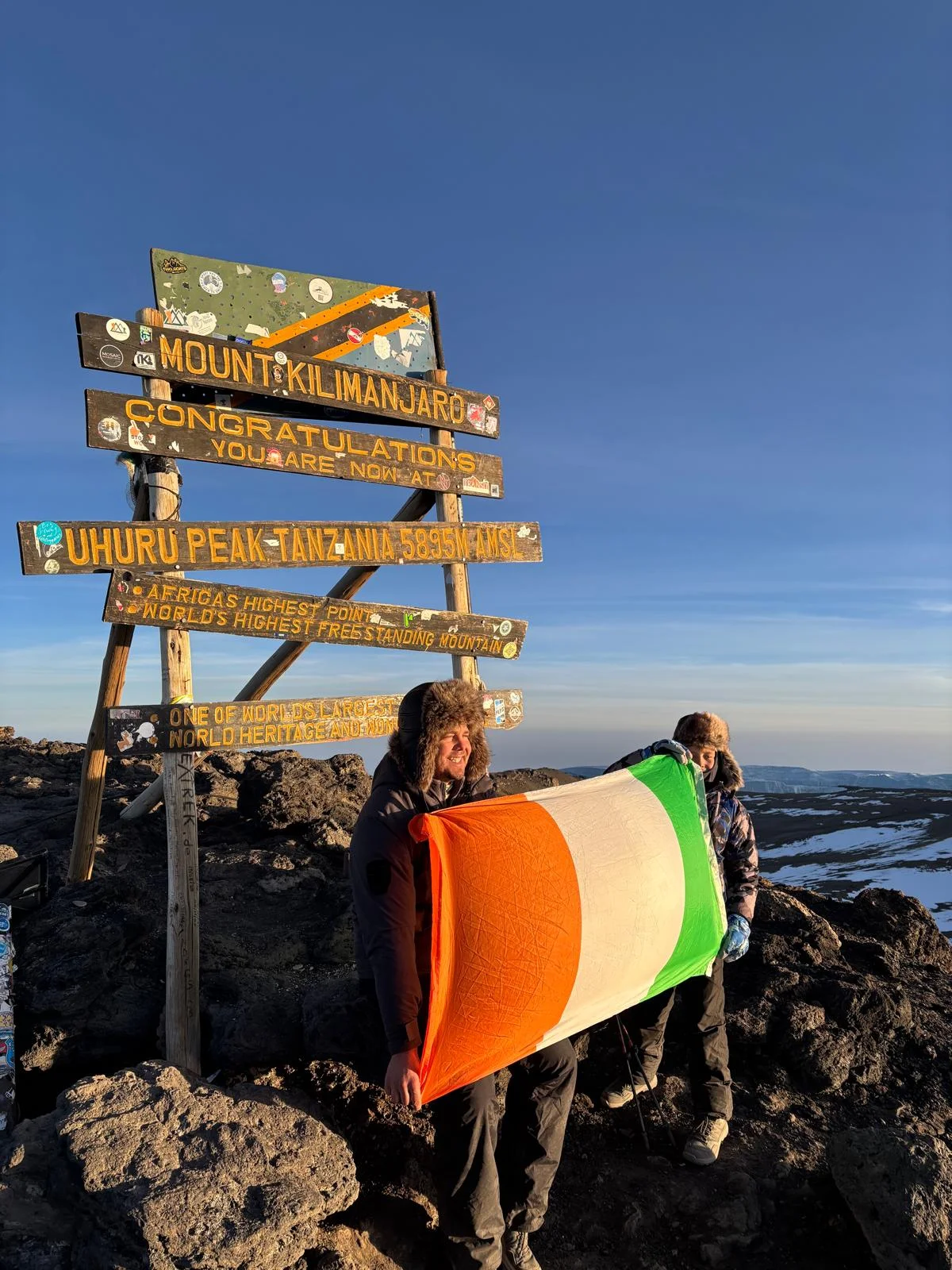 Trekkers celebrate at Uhuru Peak on Mount Kilimanjaro holding a flag beside the summit sign under clear Tanzanian skies today