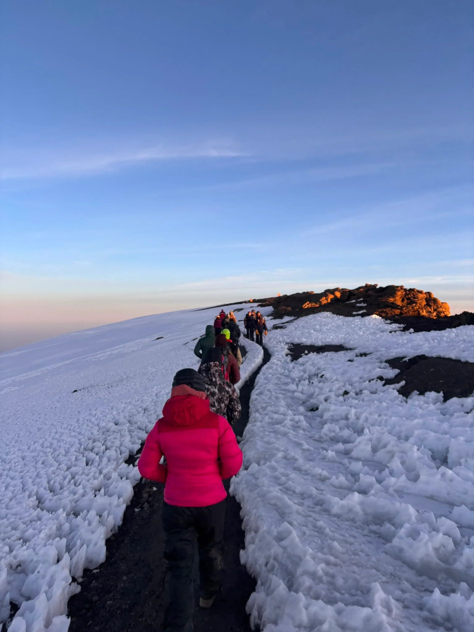 Trekkers follow a snowy trail near Mount Kilimanjaro's summit at sunrise, climbing through icy terrain under deep blue skies.