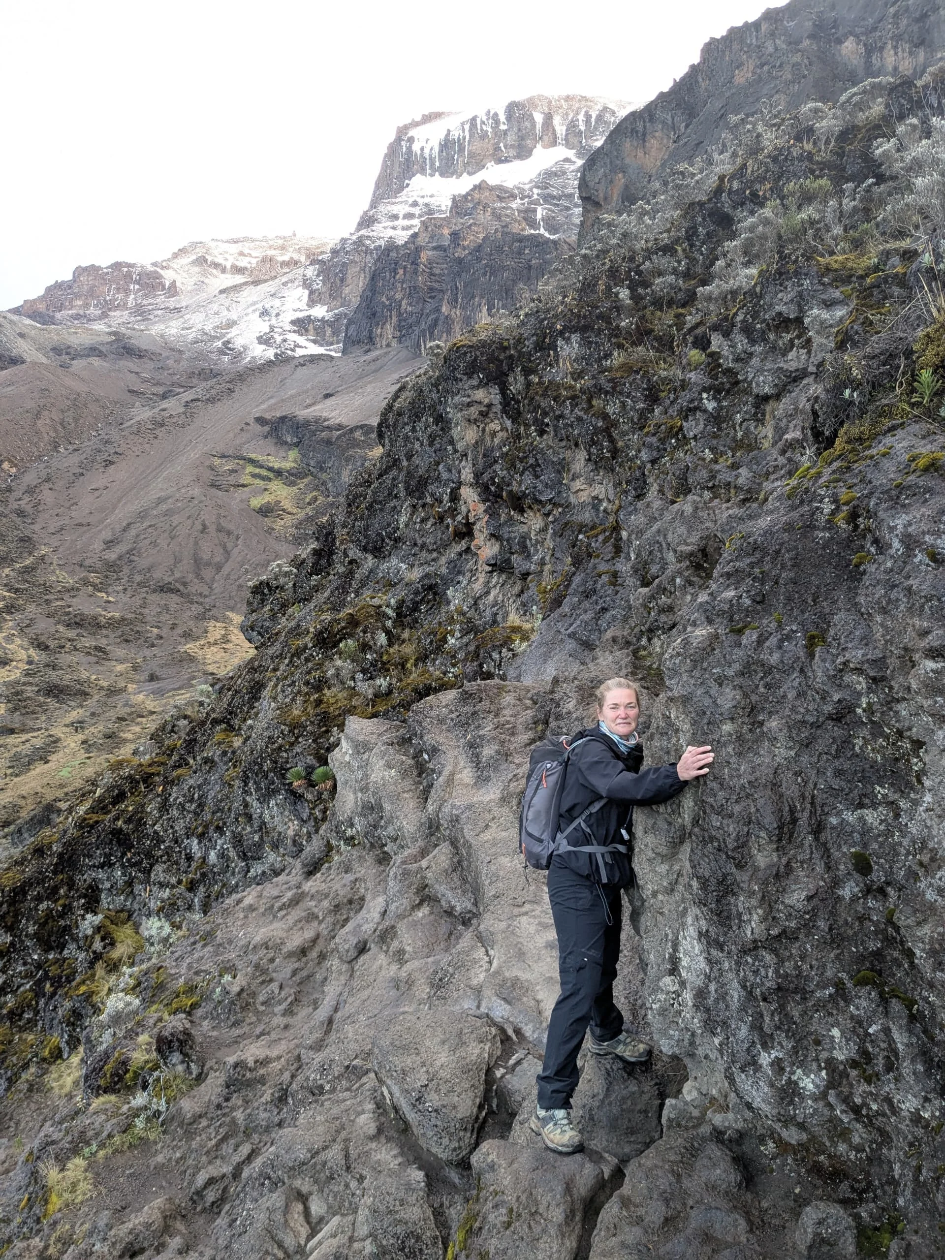 A trekker climbs rocky terrain on Mount Kilimanjaro's Lemosho Route beneath cliffs and snow-covered summit slopes in Tanzania