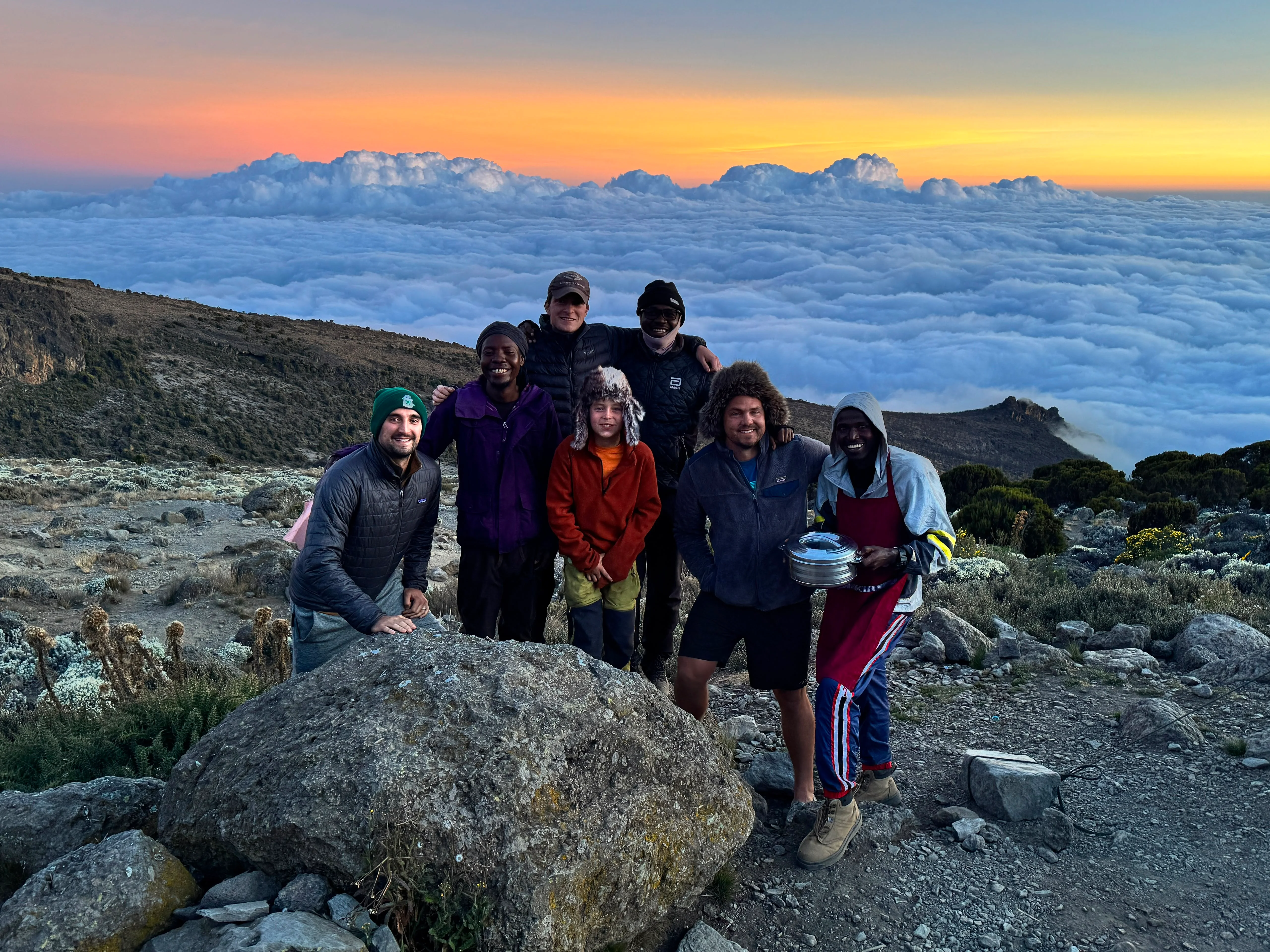 Group of Mount Kilimanjaro trekkers pose above a sea of clouds at sunset on Tanzania's scenic Northern Circuit Route at dusk.
