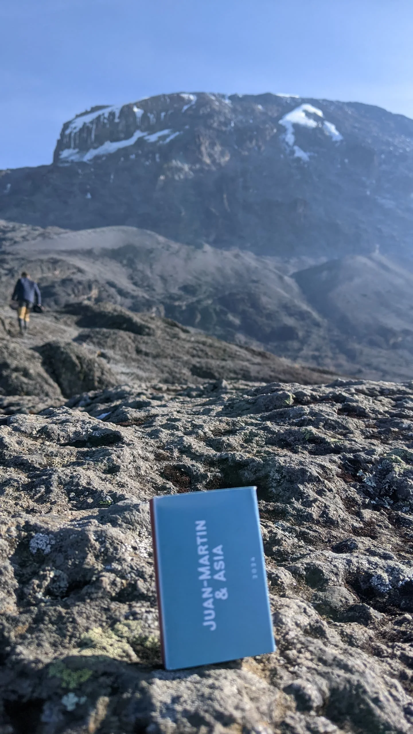 Rocky alpine terrain on Mount Kilimanjaro with a snow-dusted summit rising above the trail beneath a clear blue Tanzania sky.