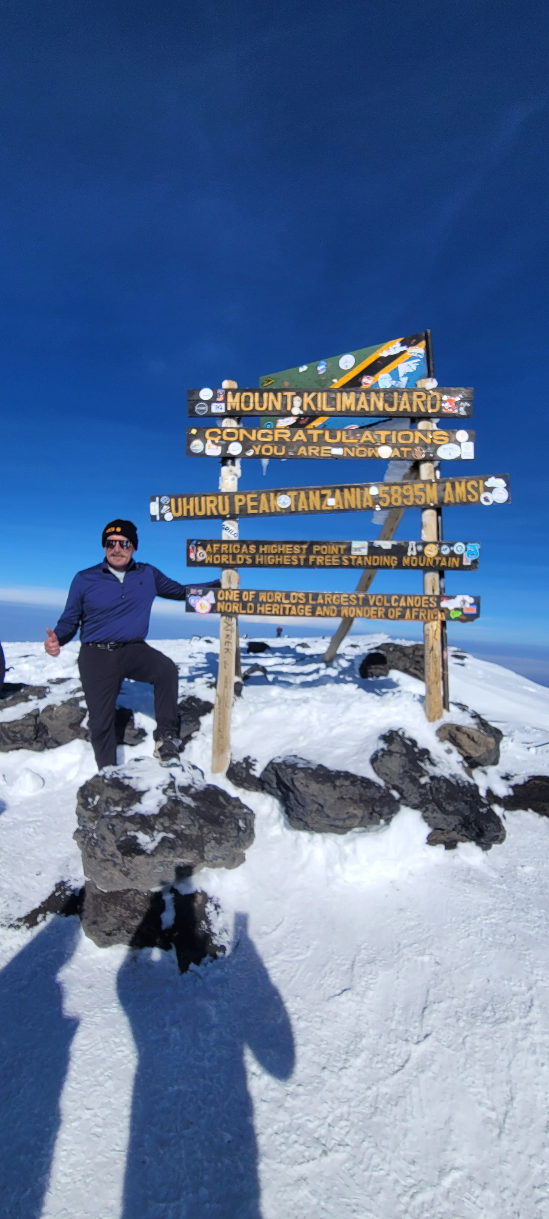 Solo climber stands at snowy Uhuru Peak on Mount Kilimanjaro beside the summit sign under deep blue Tanzanian skies above.