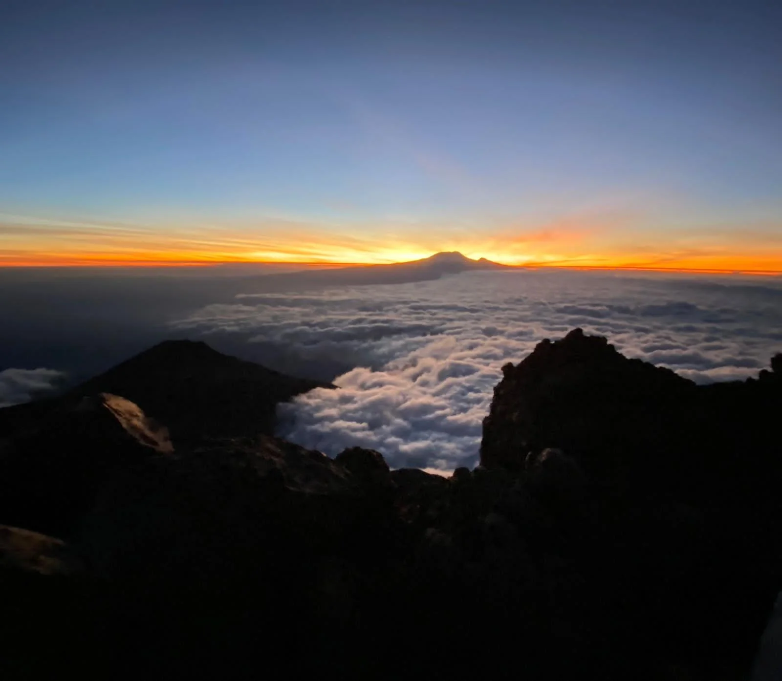 Dramatic sunrise glowing orange over dark mountain peaks and a sea of clouds on Mount Kilimanjaro.