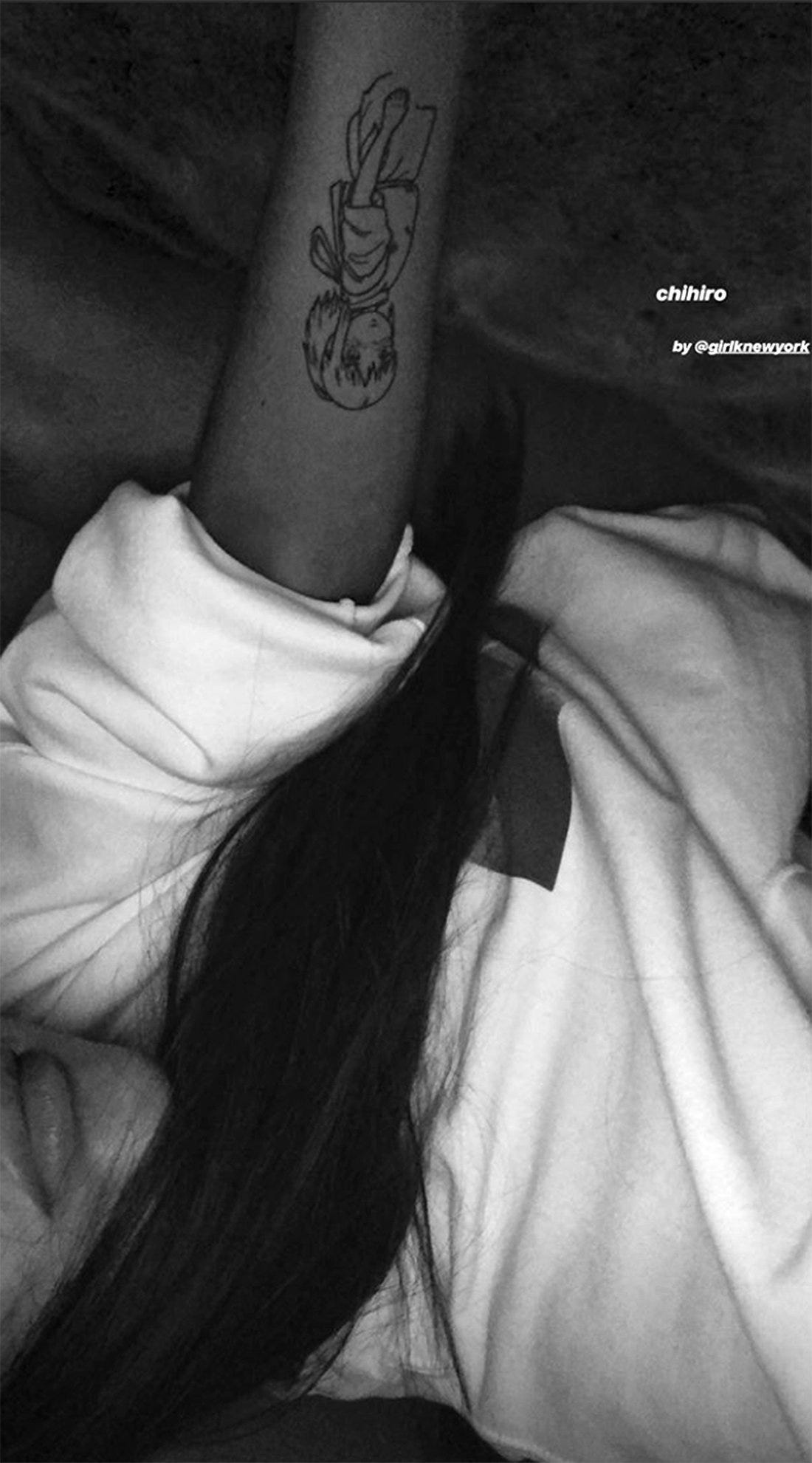 Ariana Grande raising her right forearm to show the full black line-art Chihiro portrait