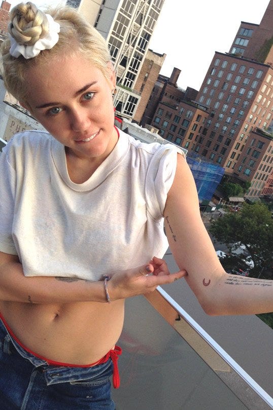 Miley Cyrus points toward the small outlined crescent moon on her left inner elbow in a rooftop photo.