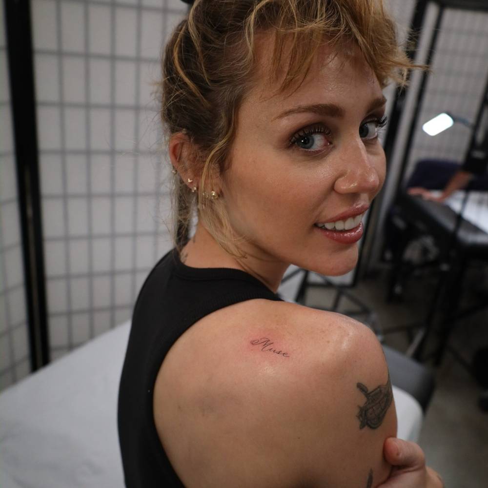 Miley Cyrus looking back over her right shoulder, showing the complete small Muse tattoo high on the shoulder blade.