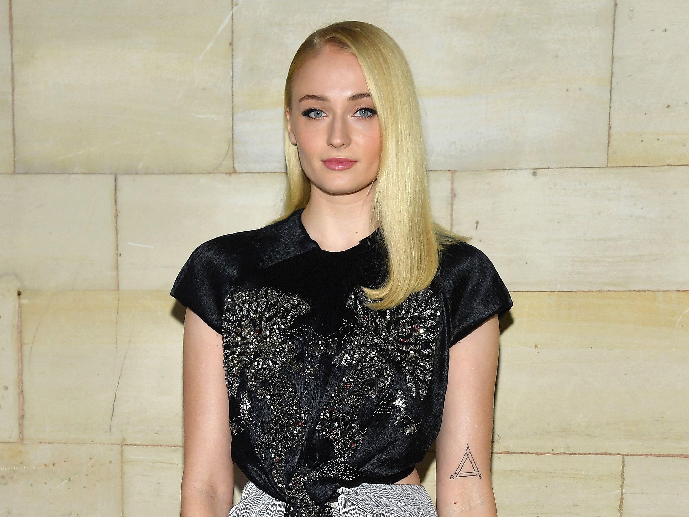 Sophie Turner wearing a black top, with the complete small triangle tattoo visible on her left upper arm.