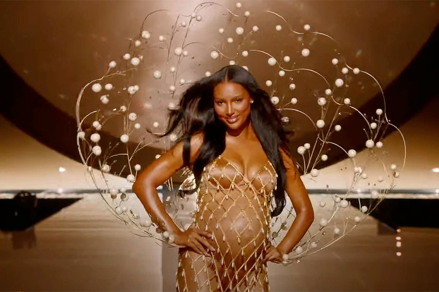 Pregnant model wearing an intricate gold mesh dress with decorative spheres at a Victoria's Secret fashion show runway