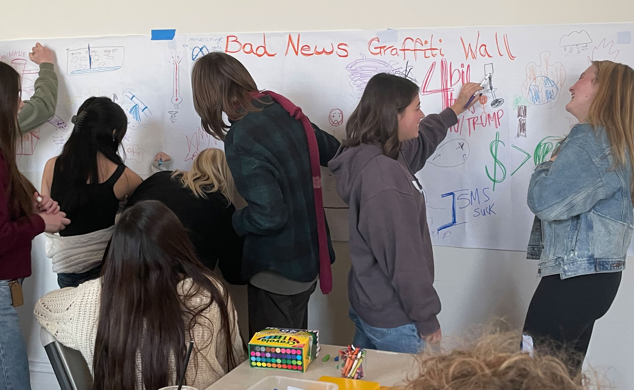 Jóvenes apiñados junto a un muro mientras dibujan en un largo trozo de papel colgado allí, en cuya parte superior puede leerse, en caligrafía grande, "Muro de Grafitis de Malas Noticias".